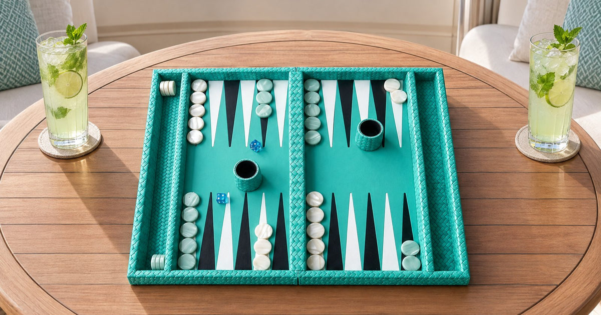 A turquoise folding leather backgammon board lies open on a yacht's teak flybridge table between two drinks, with checkers and two leather-covered dice cups in play.
