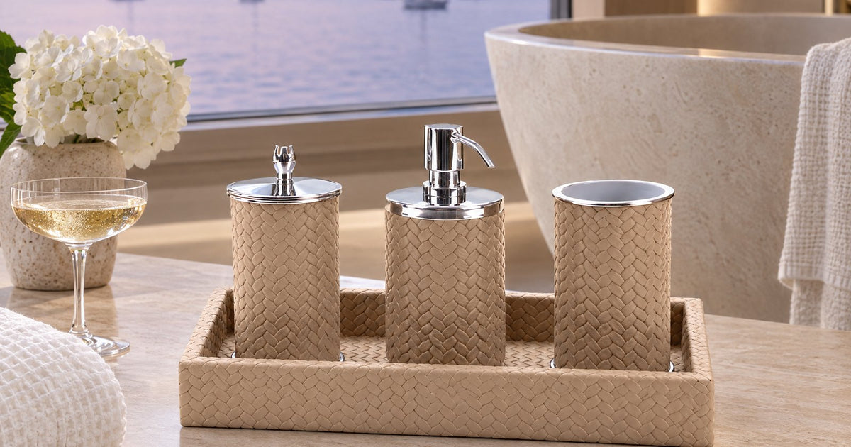 A sand-coloured four-piece bathroom set in woven-embossed leather on its tray on a stone bathroom counter, anchored sailing yachts at dusk through the window.