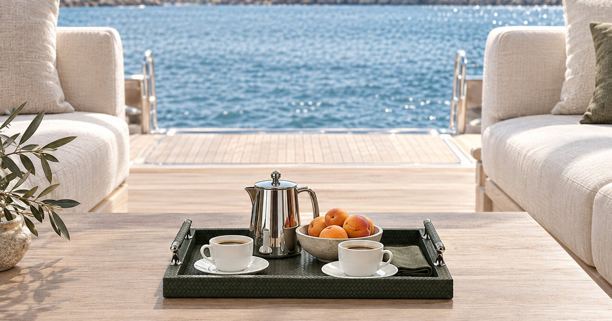 A woven-embossed leather serving tray with polished steel handles holds a coffee service and apricots on a yacht's open aft deck, the sea behind.