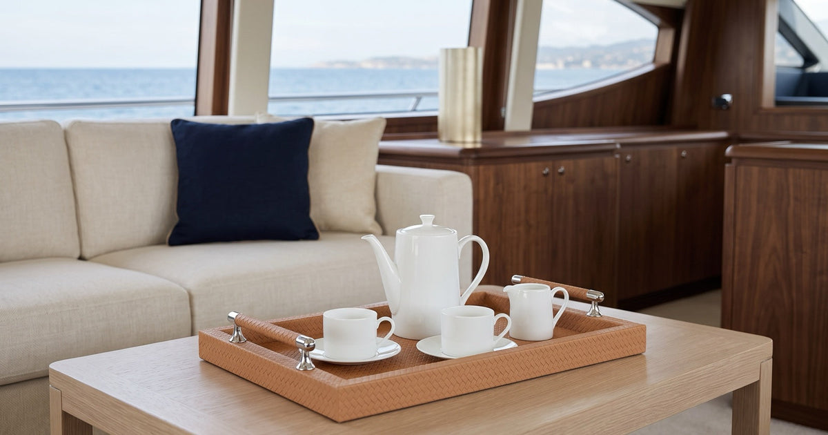 A tan woven-embossed leather serving tray with stainless steel handles carrying a coffee service on a low table in a yacht salon, the sea visible through the windows.