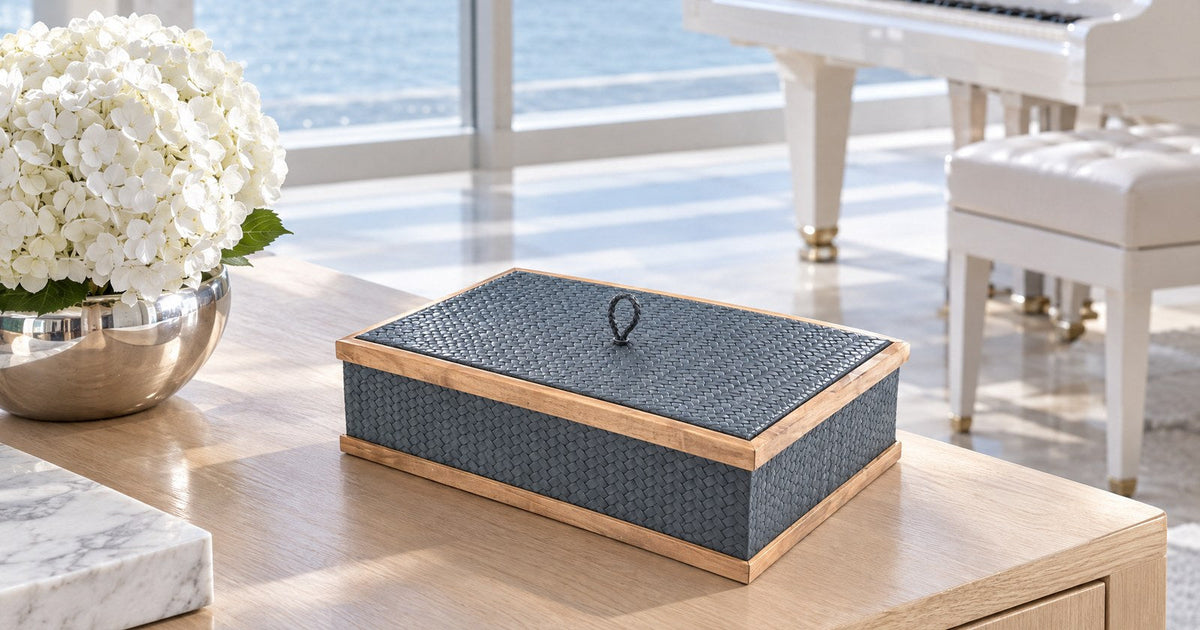A navy leather box with a woven texture, a light walnut frame and a small leather loop on its lid sits on a pale timber console in a bright salon, with a white grand piano and the sea behind.