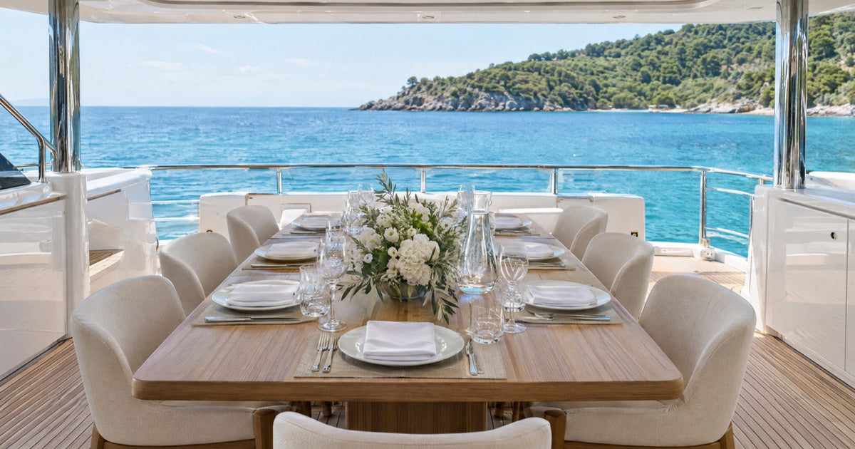 The aft deck of a motor yacht laid for lunch at anchor, with crisp white linen and glassware on the table and a calm sea behind.
