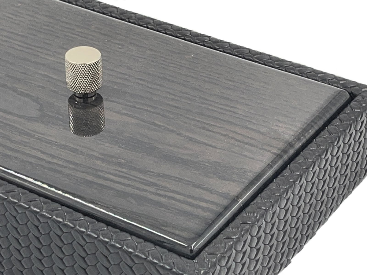Close on a corner of the 27.5 x 20 cm box, the polished wood lid edge meeting scale-embossed leather beside a knurled metal knob